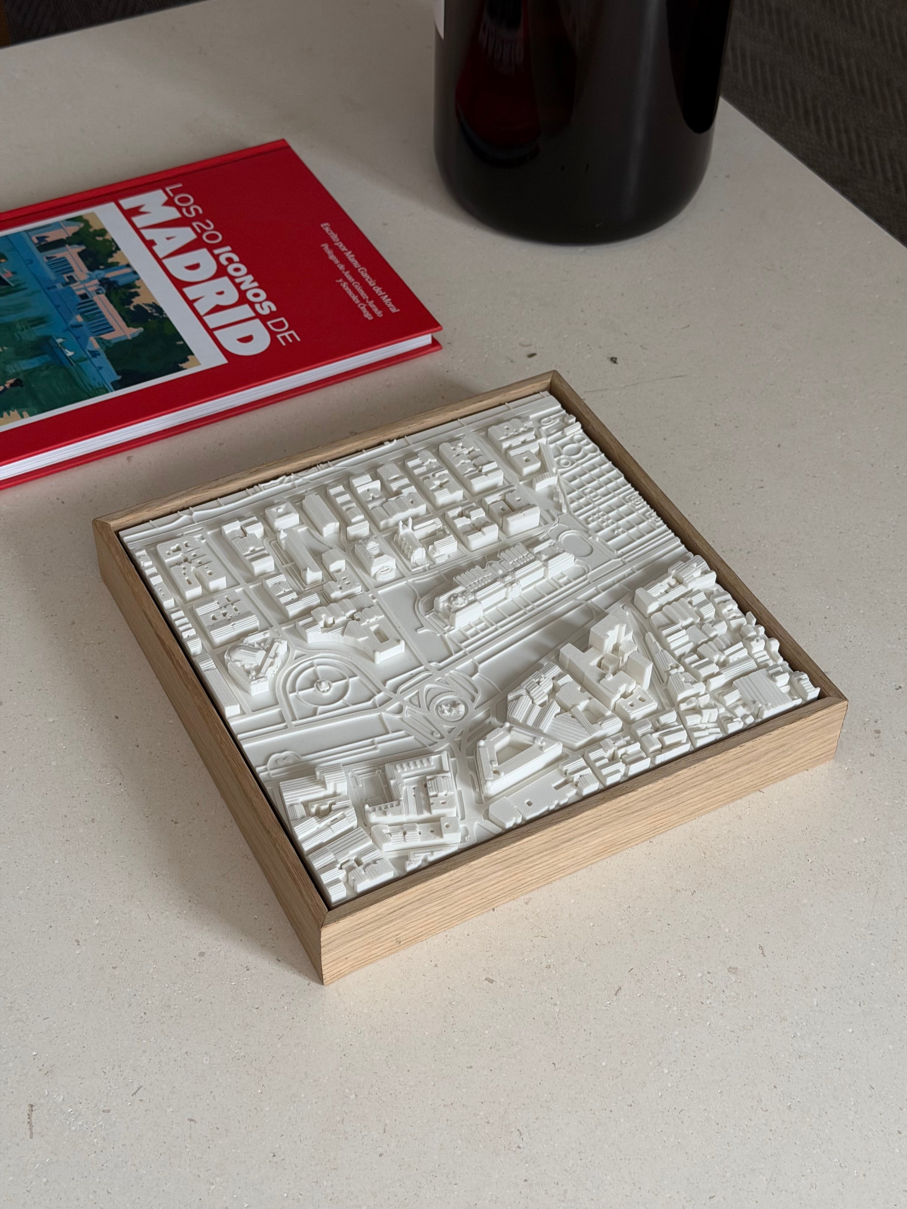 3D Madrid city map wall art in a wooden frame on a minimalist table, with guidebook nearby.