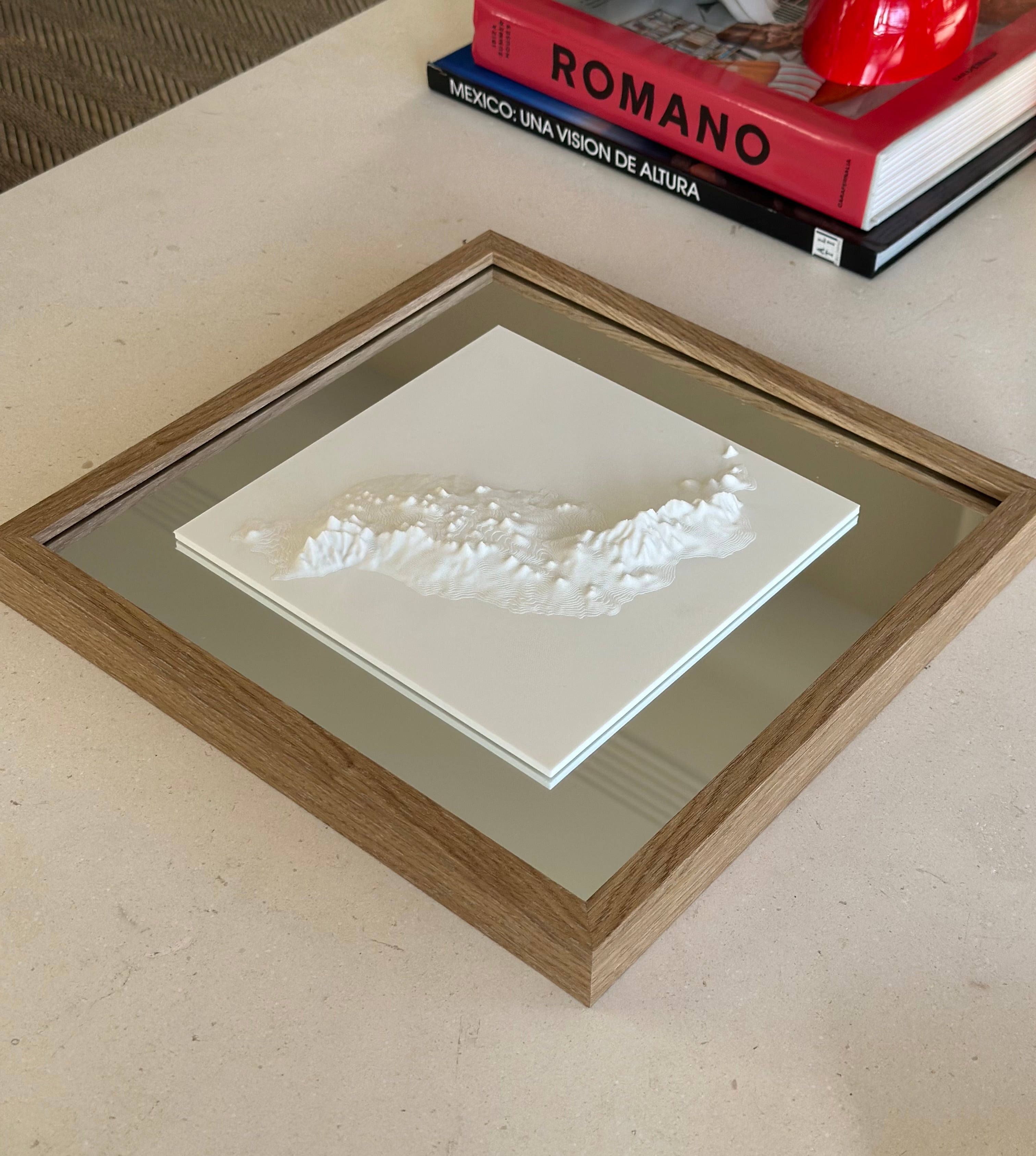Framed 3D city map artwork with white relief on a mirrored surface, on a table beside books.